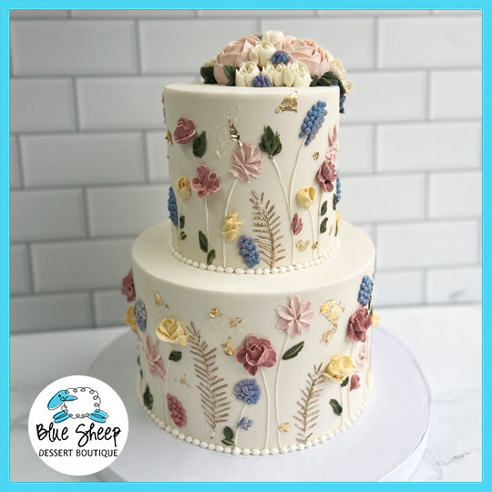 Elegant two-tiered custom cake with intricate wildflower decorations in soft pastel colors, topped with a bouquet.