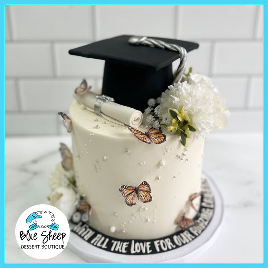 A single-tier graduation cake decorated with white flowers, delicate butterflies, and a graduation cap with diploma on top.