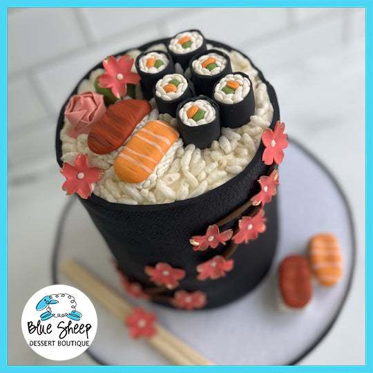 A cake designed to look like a sushi roll platter, complete with detailed fondant sushi pieces, rice, and pink floral decorations.