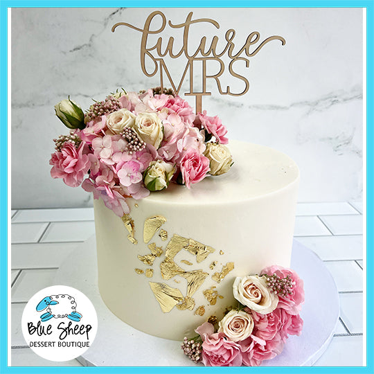 Elegant bridal shower cake with 'Future Mrs' topper, adorned with pink and white fresh flowers and gold leaf details.