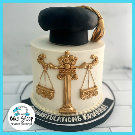 A single-tiered law-themed graduation cake with a graduation cap on top, featuring a golden Scales of Justice emblem and 'CONGRATULATIONS [Name]' on the base.