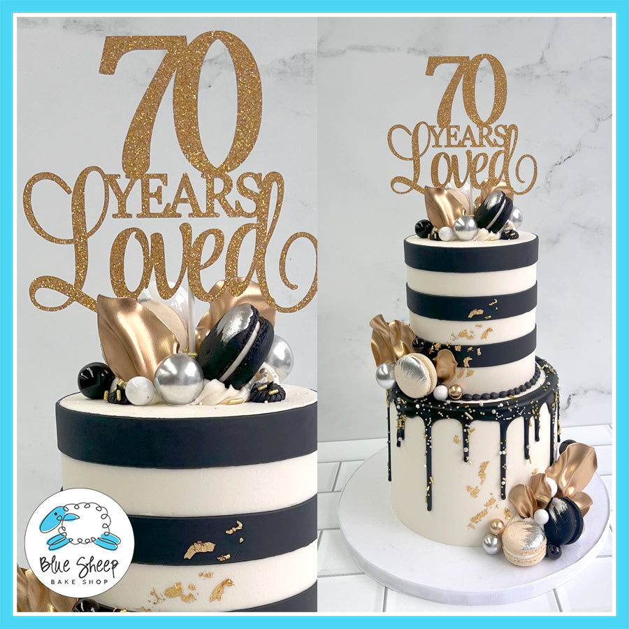 Elegant two-tiered cake with black and white stripes, gold accents, golden flowers, macarons, edible pearls, and "70 Years Loved" topper.