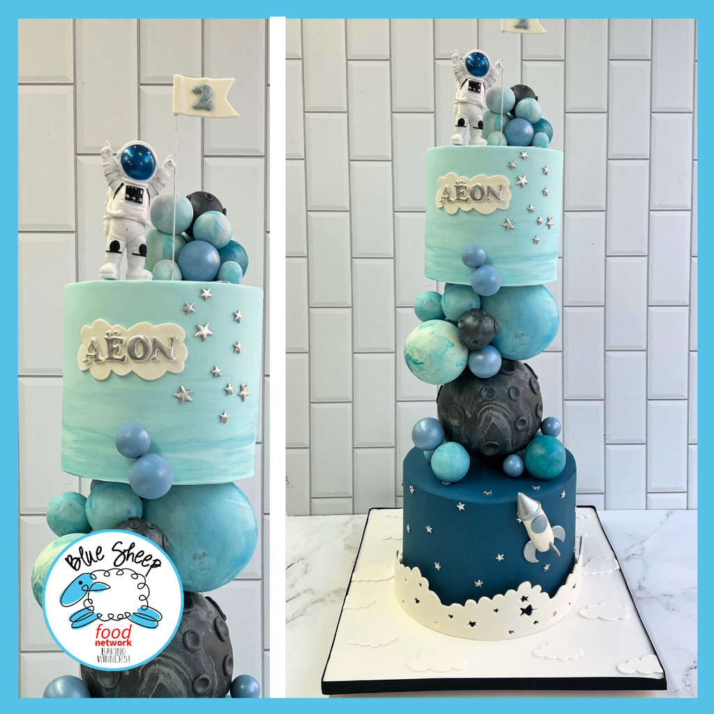 Space-themed tiered birthday cake with blue and gray planetary spheres, an astronaut topper, and custom name tag set against a hand-painted galaxy backdrop.
