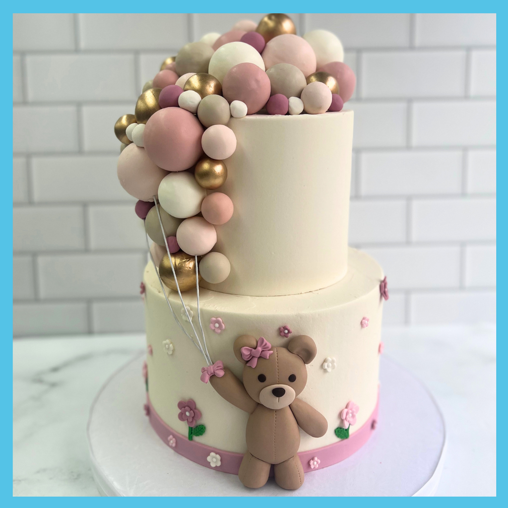 Elegant two-tiered celebration cake with pink and gold fondant balloon garland and a cute teddy bear decoration, on a white and pink floral base
