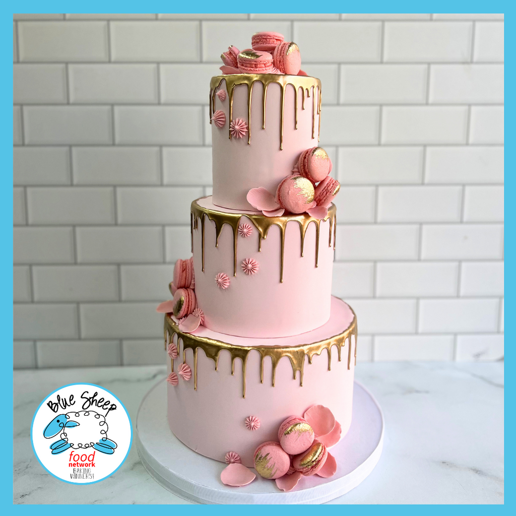 Luxurious three-tiered blush pink cake with gold drip icing, adorned with pink macarons, gold dusting, and delicate sugar work.