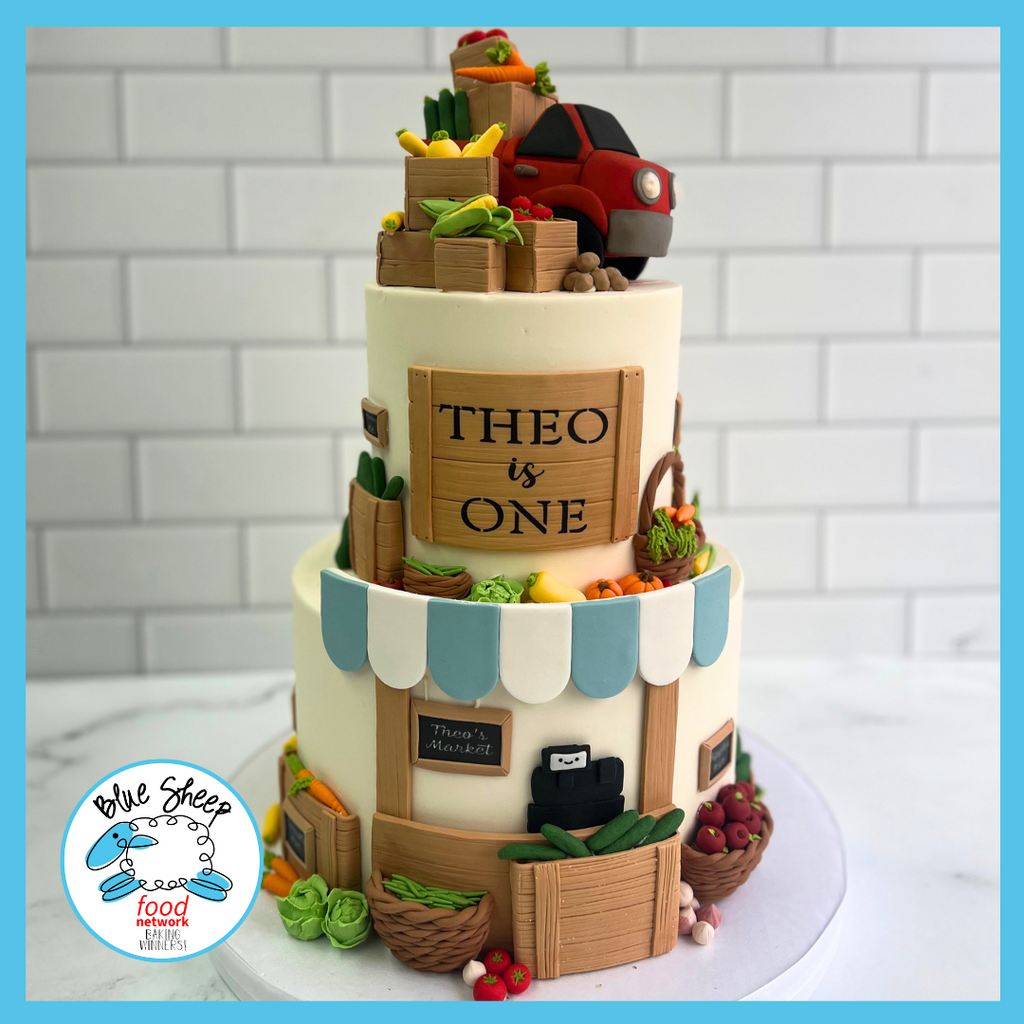 Two-tier farmer's market themed first birthday cake with fondant vegetables, wooden crate designs, and a red car topper, customized with 'Theo is One'