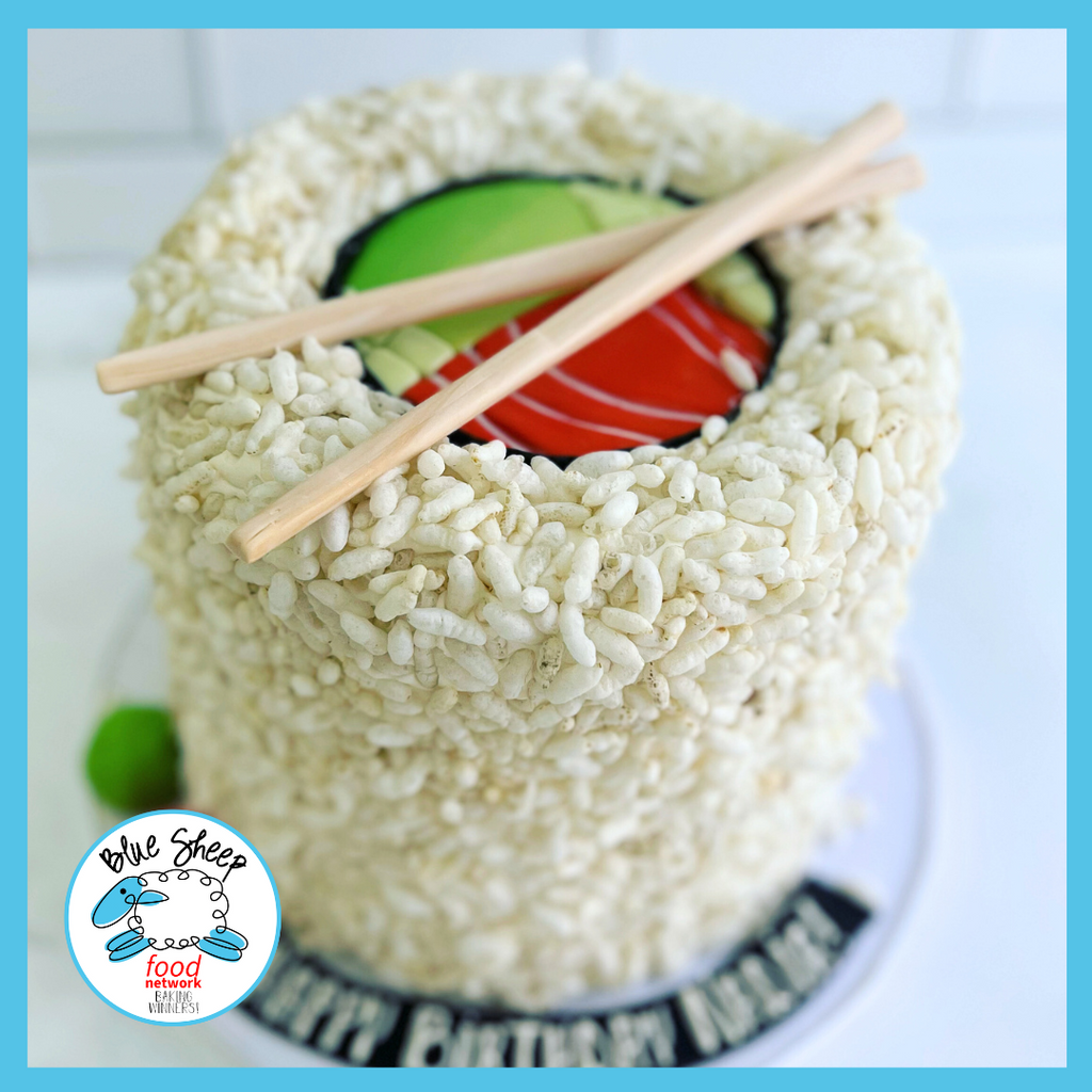 Novelty sushi roll cake decorated with puffed rice to mimic sushi rice, edible fondant seaweed, colorful fillings, and a pair of chopsticks on top, presented on a white background.