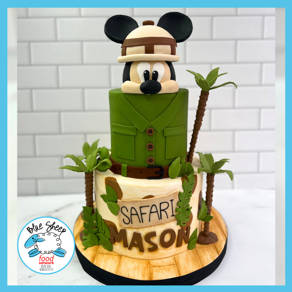 Custom jungle explorer themed birthday cake with a fondant cartoon character in safari outfit, surrounded by fondant palm trees and leaves, with 'Safari Mason' text.