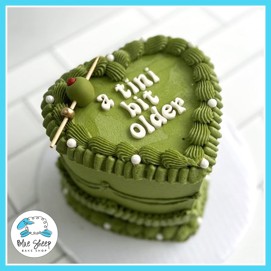 Green heart-shaped birthday cake with martini-inspired design and fondant olive topper, reading 'A Tini Bit Older