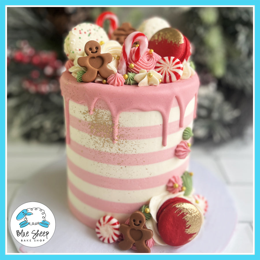 Christmas-themed cake with pink and white stripes, pink drip, gingerbread cookies, candy canes, macarons, and festive buttercream decorations.