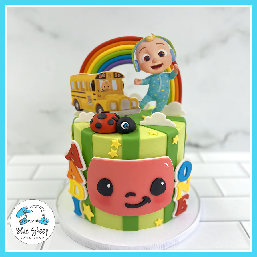 Cocomelon-themed first birthday cake with Cocomelon face, rainbow, yellow school bus, baby JJ, and colorful decorations.