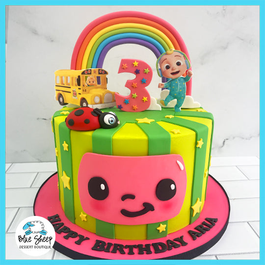 Cocomelon themed custom birthday cake with rainbow, Cocomelon characters, school bus, ladybug, and personalized message.