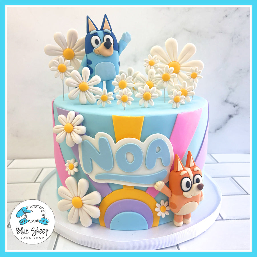 Custom Bluey-themed birthday cake with fondant Bluey and Bingo figurines, daisies, and rainbow decorations by Blue Sheep Bake Shop in Somerville, NJ.