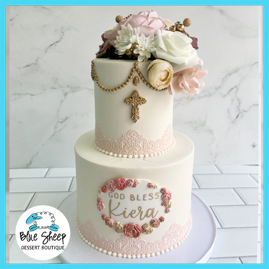 A two-tiered First Holy Communion cake with lace detailing, rose adornments, and a 'God Bless Kiera' inscription.