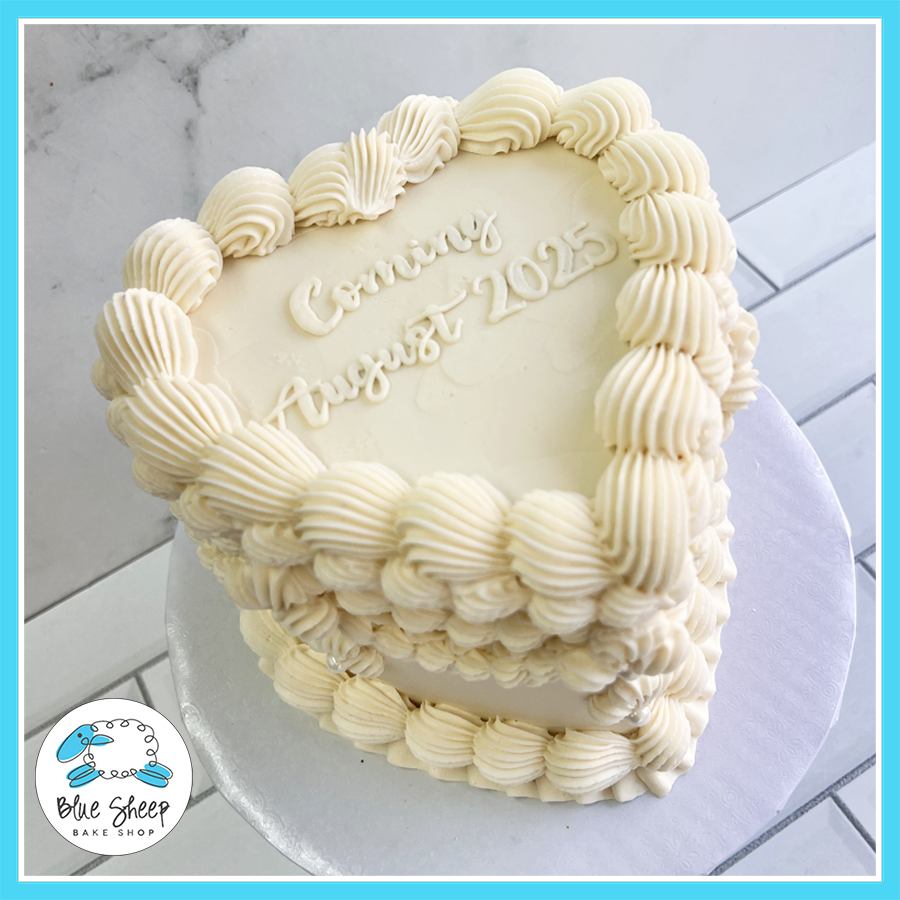 Heart-shaped baby announcement cake with piped vintage buttercream, elegant white frosting, and a custom message, available at Blue Sheep Bake Shop in Somerville, NJ

