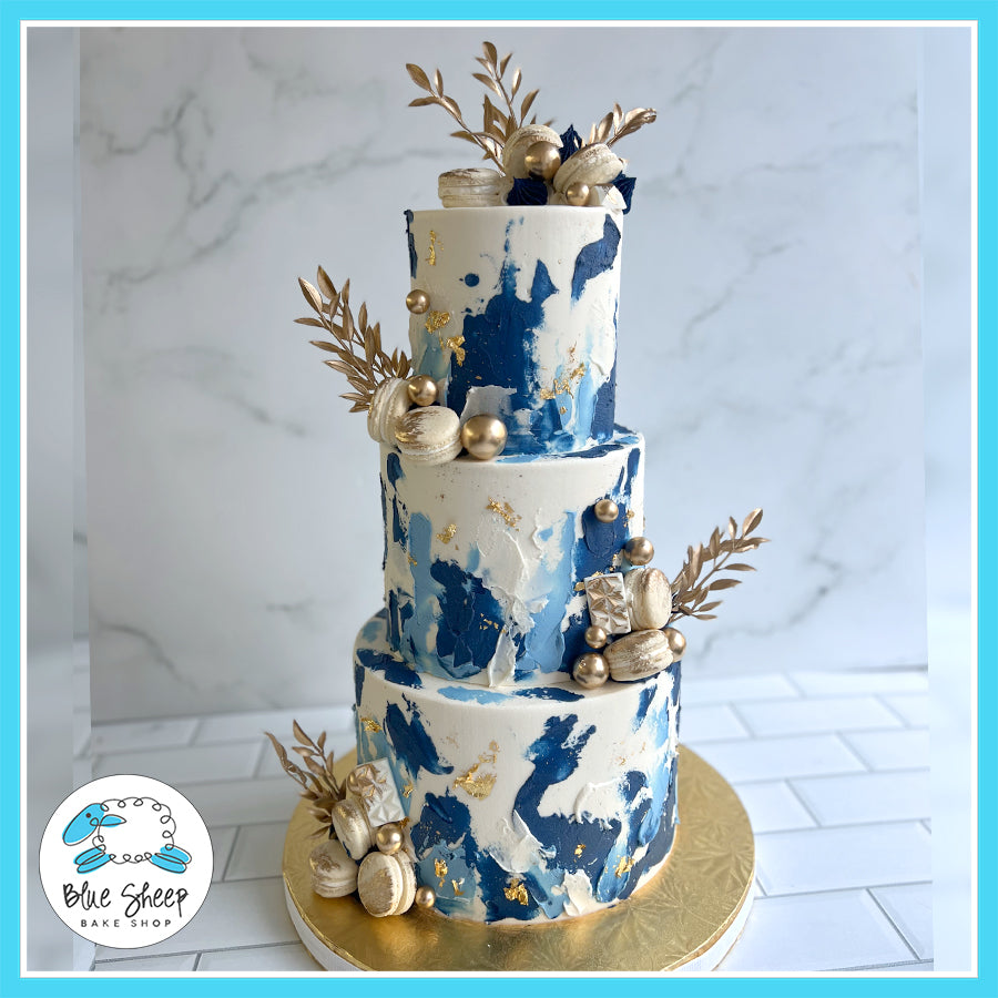 Three-tier navy blue and white abstract design cake with gold accents, macarons, and metallic decorations