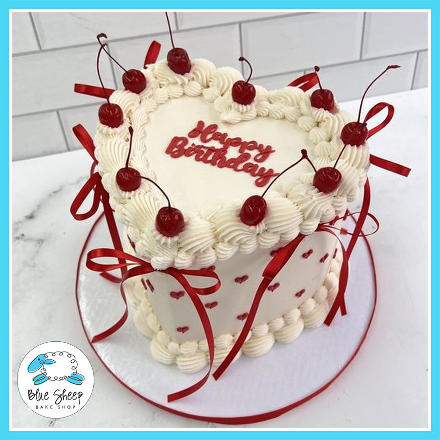 Heart-shaped birthday cake with vanilla buttercream, maraschino cherries, red ribbon accents, and red heart decorations.