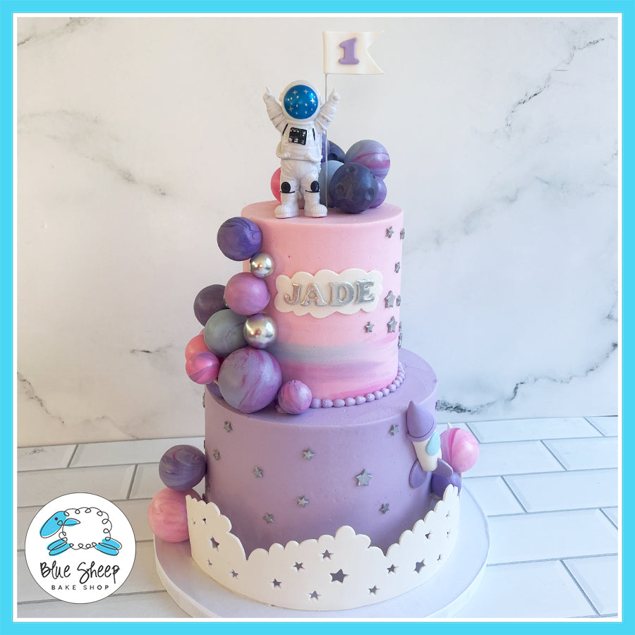 Two-tier space-themed cake with astronaut topper, galaxy spheres, and silver star decorations by Blue Sheep Bake Shop.