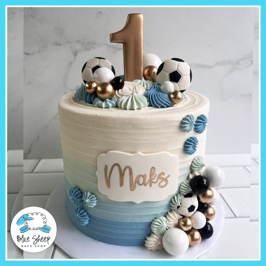 Elegant soccer-themed birthday cake with ombré blue and white buttercream, edible soccer ball decorations, gold accents, and a personalized name plaque.