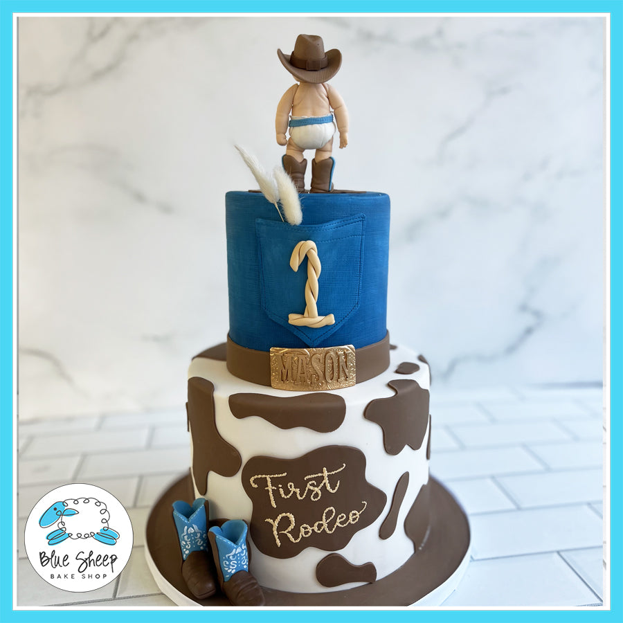 First Rodeo" cowboy-themed birthday cake with cowhide patterns, denim accents, a cowboy figurine, and mini boots by Blue Sheep Bake Shop in Somerville, NJ.