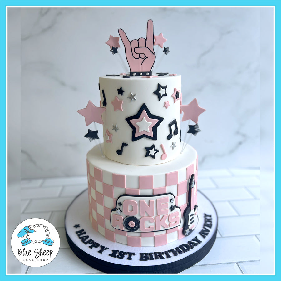 Two-tier pink and white rock star-themed birthday cake with stars, musical notes, and "One Rocks" sign