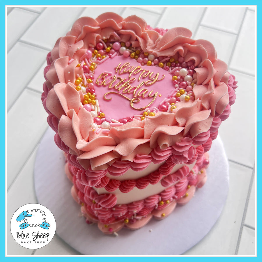 Pink heart-shaped birthday cake with vintage buttercream ruffles, pearl sprinkles, and a gold "Happy Birthday" topper, available at Blue Sheep Bake Shop in Somerville, NJ