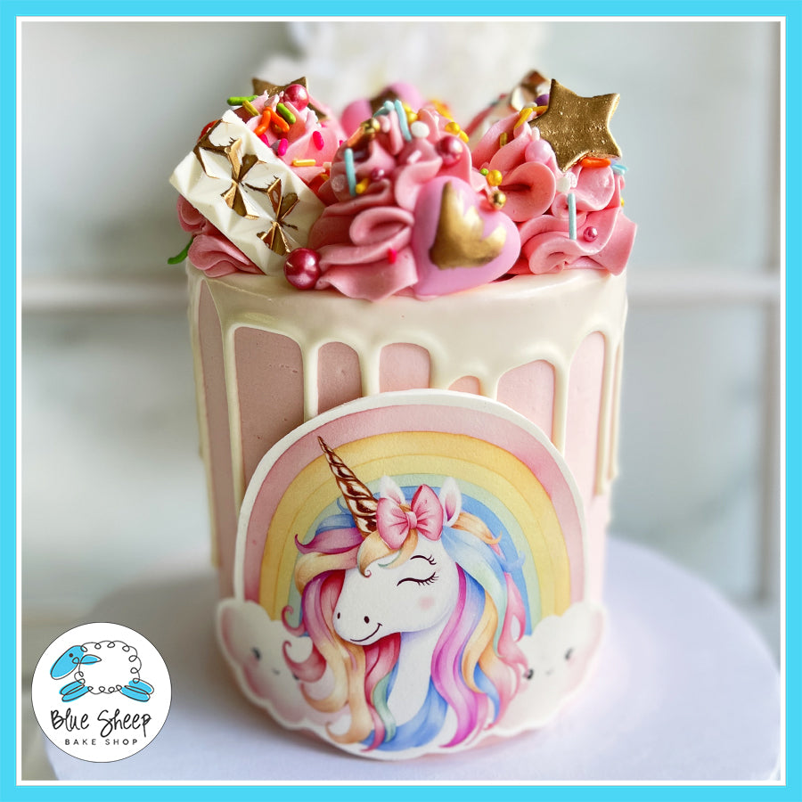 Pink unicorn-themed drip cake with a rainbow plaque, buttercream swirls, and gold accents by Blue Sheep Bake Shop.