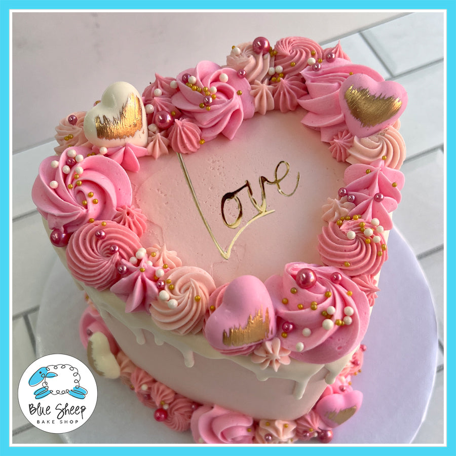 Pink heart-shaped valentine's cake with gold lettering, buttercream florals, and chocolate heart toppers from Blue Sheep Bake Shop in Somerville, NJ