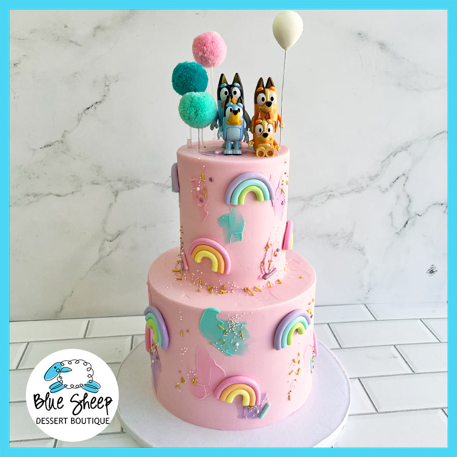 Custom two-tiered birthday cake with pink fondant, colorful rainbows, and character toppers, created by Blue Sheep Dessert Boutique bakery