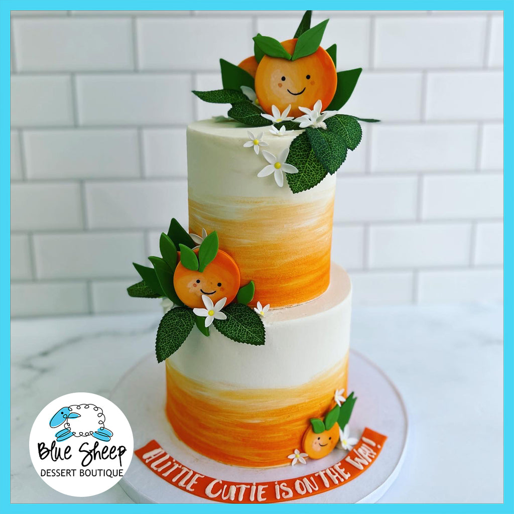 Two-tier custom baby shower cake with an 'A Little Cutie is on the Way' theme, decorated with fondant orange slices, smiling citrus characters, and white blossoms against a gradient orange background.
