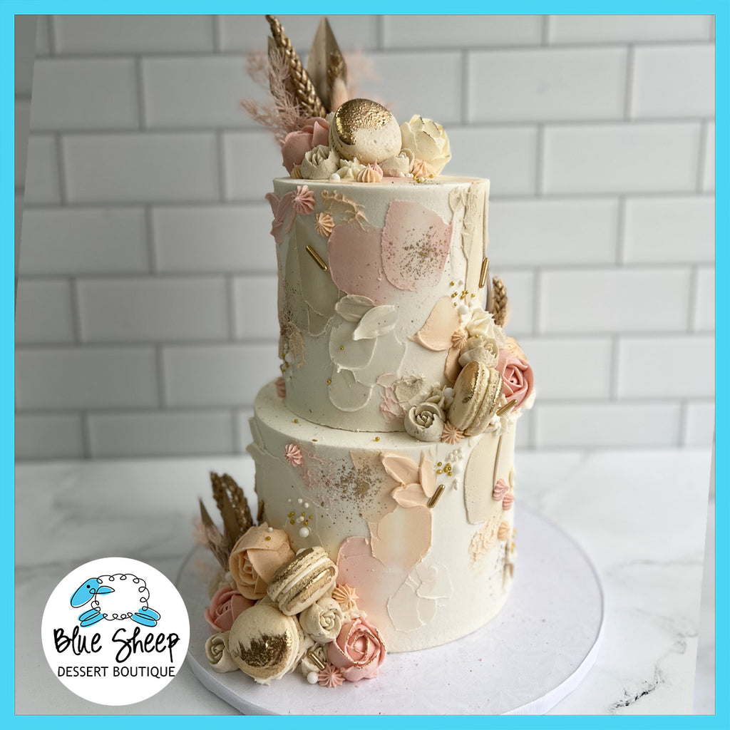 Elegant two-tier custom birthday cake in blush and cream tones with gold accents, edible roses, macarons, and hand-painted details, presented on a white background.