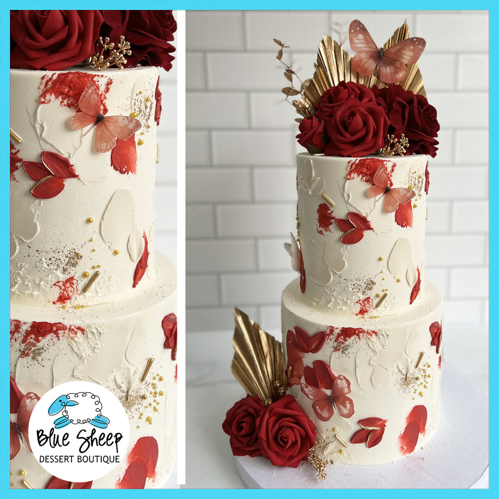 Elegant custom birthday cake with deep red roses, gold leaf accents, and butterfly decorations, set against a white and rouge watercolor backdrop.